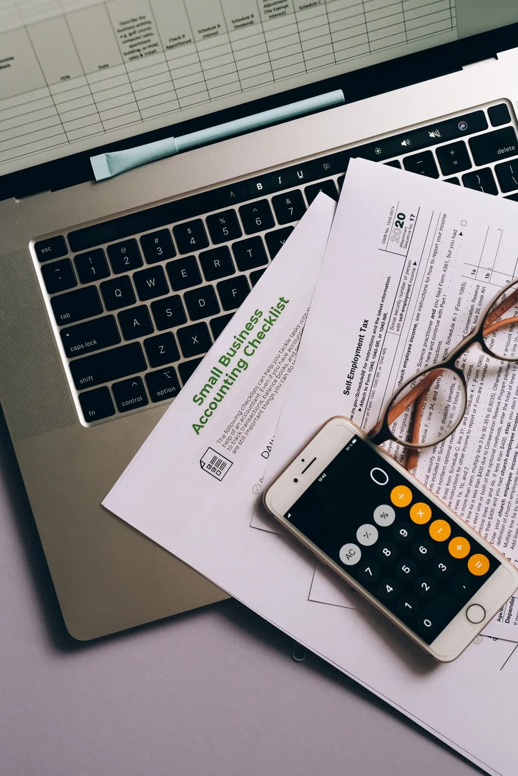 A small business accounting checklist with a phone calculator and glasses on a laptop