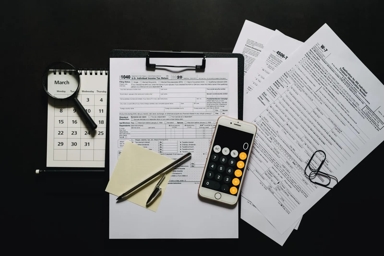 Tax forms, a calendar, a calculator and a clipboard laid out on a dark desk