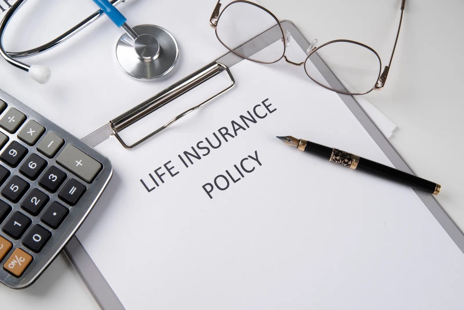 A life insurance policy on a clipboard with a pen, glasses, stethoscope and calculator